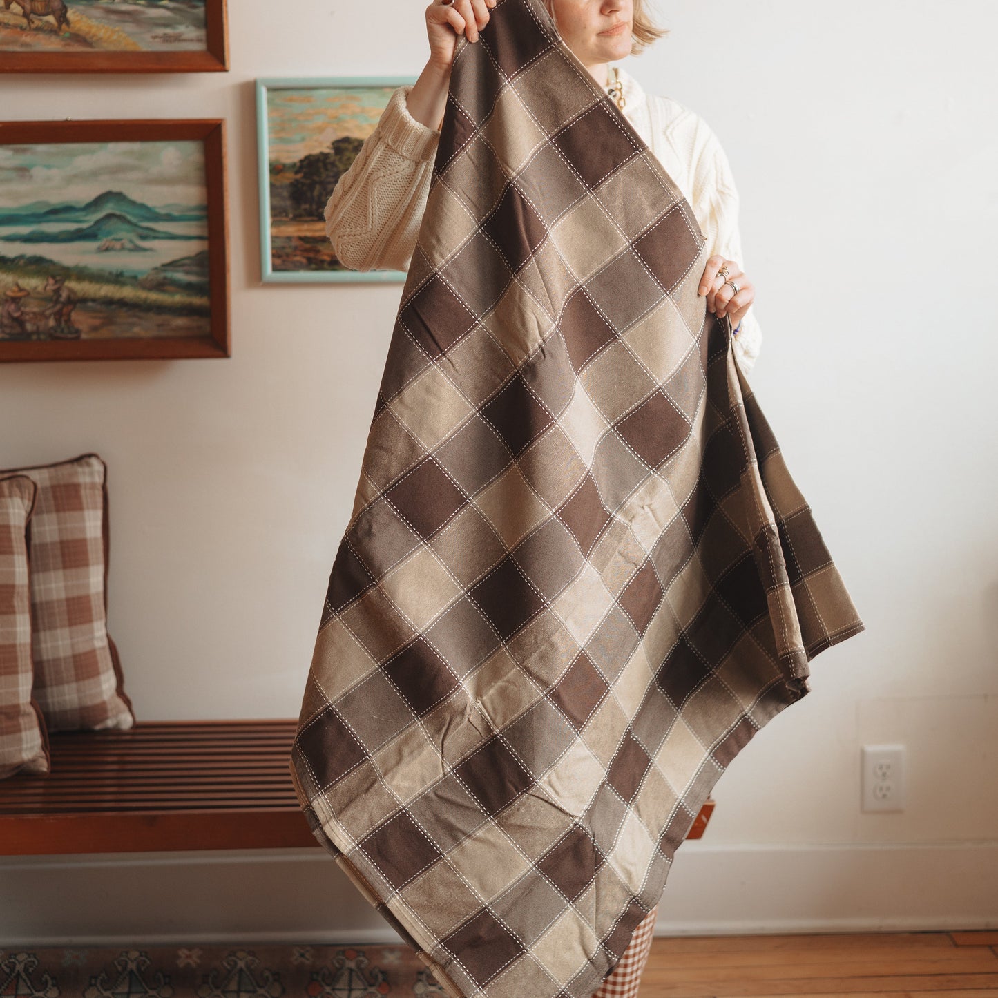 Person holding a plaid blanket in a room with wooden floors and framed pictures on the wall.